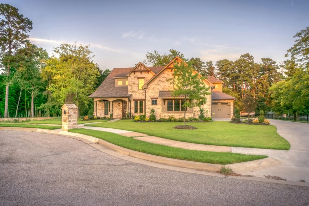 Elegant stone family home with manicured lawn, trees, and driveway on a sunny day.